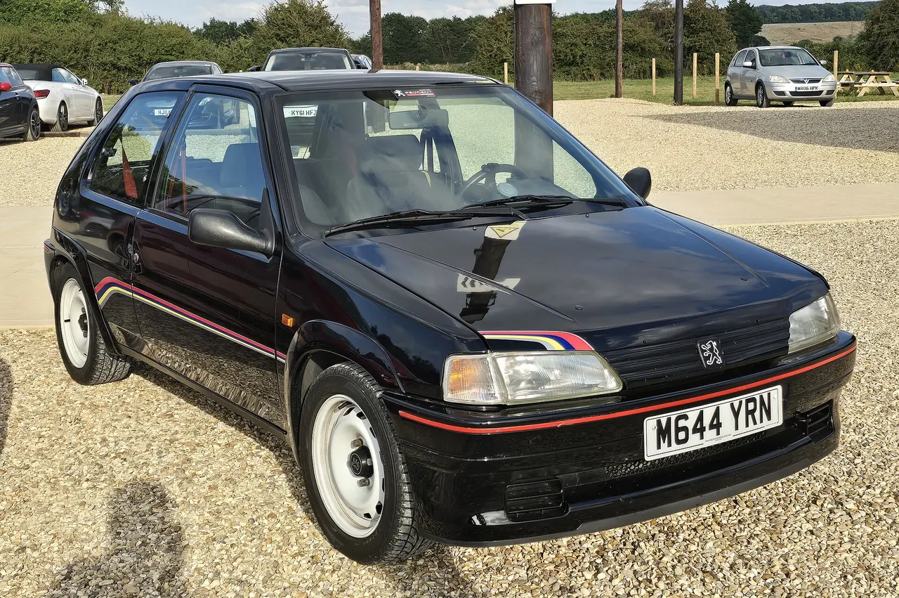 Black Peugeot 106 Rallye with a coloured side stripe, parked on gravel.